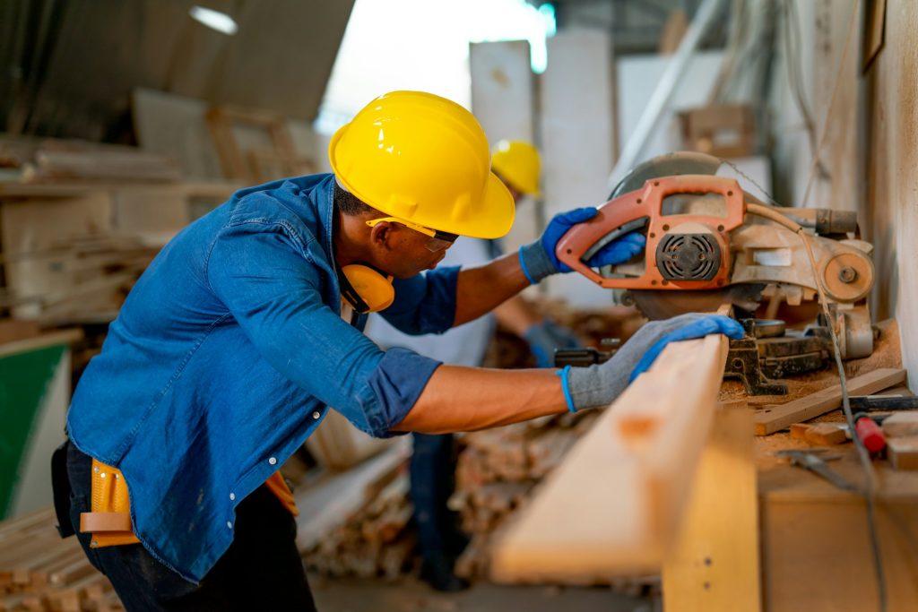 African American carpenter man use electrical saw to cut timber in factory workplace